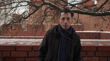 Kışın açık havada dikilen ve doğrudan kameraya bakan bir adamın portresi. Karla kaplı tarihi bir şehir duvarının arka planına karşı sakin ve tarafsız bir ifadesi var..