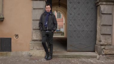 Tarihi bir şehir duvarına yaslanmış günlük kıyafetler giymiş bir adamın uzun bir portresi. Kendine güvenerek poz veriyor, klasik bir Avrupa ortamında kentsel bir görüntü sergiliyor..