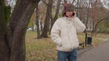 Sıcak ceketli ve şapkalı bir adam akıllı telefonuyla sohbet ediyor. Bir sonbahar şehir parkında duruyor, muhtemelen bir arkadaşını ya da randevusunu bekliyor, bağlı kalıyor ve dışarıda konuşuyor..