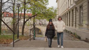 Genç bir çift, sonbaharda şehir gezintisi sırasında birlikte taş bir merdivenden yukarı çıkıyorlar. Arka planda klasik mimari ve sonbahar yapraklarıyla bir Avrupa şehrinde yürüyorlar..