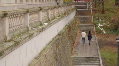 Genç bir erkek ve kadın sonbahar parkında çok katlı bir merdivene çıkıyorlar. Yolları, üzerinde klasik bir balustrade bulunan devasa bir taş istinat duvarı boyunca uzanıyor..