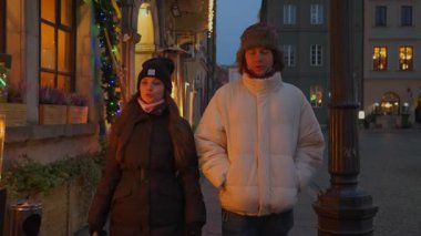 Genç çift Noel tatilinde güzel dekore edilmiş bir caddede yürüyor. Açık hava ateş ısıtıcılarının yanından geçiyorlar, akşam buluşmalarında sıcak ve büyülü festival atmosferinin tadını çıkarıyorlar.