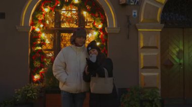 Akşam randevusu olan mutlu genç bir çift akıllı telefonuyla selfie çekmek için durur. Güzel bir Noel süslemesi önünde birlikte gülümseyip poz verirler, romantik bir anı yakalarlar..