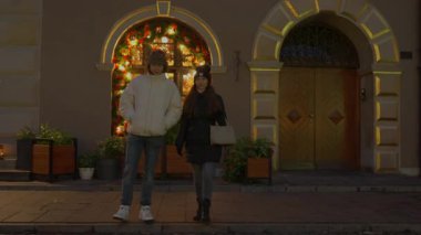 Bir adam ve bir kadın gece şehir kaldırımında birlikte yürürler. Noel tatili için ışık ve çelenklerle süslenmiş kemerli pencereli bir binanın önünden geçiyorlar..
