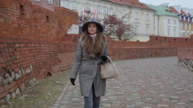 Varşova 'nın Eski Kasabası' nın kaldırım taşı caddelerinde paltolu ve şapkalı şık genç bir kadın yürüyor. Avrupa 'da turizm, seyahat ve şehir tatili kavramı.