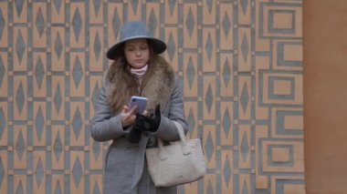 Paltolu ve şapkalı şık bir kadın, dışarıda dikilirken akıllı telefonunu iletişim için kullanıyor. Modern yaşam tarzı, iş ve teknoloji kavramı.