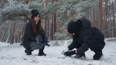 A mother and child are making a small snowman in a winter forest. Crouching together, they work as a team, shaping and forming the snowballs to stack them up. A creative family activity.
