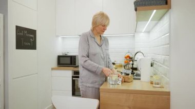 Pijamalı yaşlı bir kadın günlük rutiniyle sağlığını koruyor, ilaç ya da takviye alıyor. Su toplayan bir paketten hap alır ve bir bardak su içer..