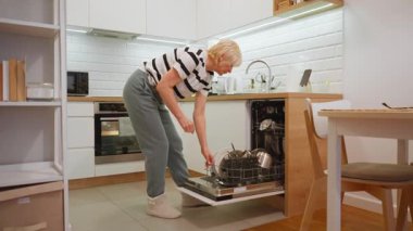 Yaşlı bir kadın bulaşık makinesini kendi mutfağında temiz mutfak gereçleriyle boşaltıyor. Tabakları ve tencereleri dolaba koyup düzenli bir ev için günlük ev işleri yapıyor..