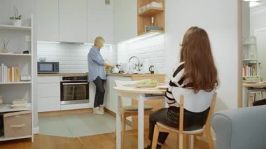 Üst düzey bir anne, ev mutfağında kızına sıcak bir içecek hazırlar. Yetişkin bir kız, ailesi bir çaydanlıktan kaynar su dökerken yemek masasında bekler..