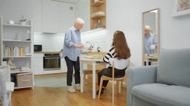 Misafirperver bir kıdemli anne yemek servisi yapıyor ve yetişkin kızı için masayı hazırlıyor. Bir kadın mutfaktan yemek ve çaydanlık getirir, aile yemeği ya da evde öğle yemeği hazırlar..