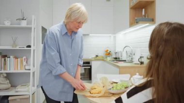 Yaşlı bir büyükanne mutfak masasında ergen torununa taze pişmiş hamur işi ikram eder. Mutlu bir aile yemeğinin tadını çıkarıyor, evde bakım ve kaynaşma anı..
