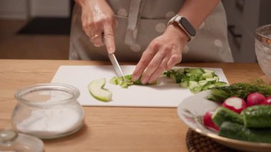 Bir ev hanımı beyaz bir kesme tahtasında taze yeşil salatalık doğruyor. Sağlıklı bir yaz salatası için sebze doğrama işlemi. Evde hafif bir vejetaryen yemeği hazırlıyorum..