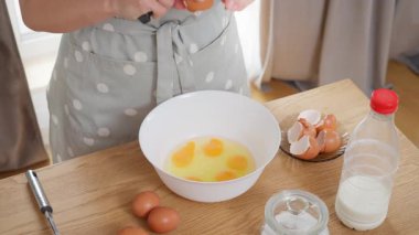 Bir kadın kahvaltı hazırlıyor, yanında çırpıcıyla taze tavuk yumurtalarını beyaz bir kâseye kırıyor. Ev yapımı bir yemeğin pişirme süreci, mutfakta protein zengini bir yemek tarifi..