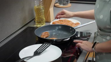 Bir kadın terbiye edilmiş tavuk göğsünü kızgın yağlı bir tavada kızartıyor. İndüksiyon sobasında tavuk eti pişirme işlemi, ev yapımı akşam yemeği için fileto parçaları yakma..
