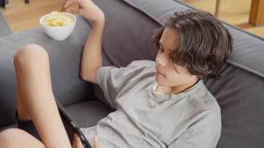 Bir çocuk evde çip yerken tablet bilgisayarının ekranı tarafından tamamen özümsenir. Ekran zamanı bağımlılığı, dijital bağımlılık ve modern çocukluk eğlence aktiviteleri kavramı.