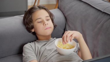 Bir çocuk beyaz bir kaseden zevk alarak çıtır patates cipsi yiyor, kanepede tabletle dinlenirken lezzetli bir aperatifin tadını çıkarıyor. Basit bir zevk ve doyurucu bir açlık anı..