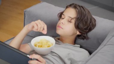 Sıkılmış bir çocuk kanepede yatarken düşüncesizce abur cubur yiyor ve dijital tablet kullanıyor. Yalnızlığın tasviri, zaman kaybı ve motivasyon eksikliği..