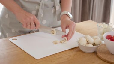 Beyaz tahtada bıçakla taze şampiyon mantarlarını doğrayan kadın elleri. Önlüklü bir kadın ev yapımı vejetaryen yemeği pişiriyor. Mutfakta sağlıklı yemek malzemeleri hazırlıyor..