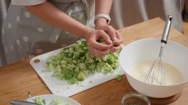 Bir kadının elleri, beyaz bir kesme tahtasından taze kesilmiş yeşil brokoli çiçeklerini mutfaktaki büyük beyaz bir kaseye aktararak yemek için gerekli malzemeleri hazırlıyor..