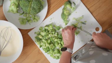 Bir kadının elleri titizlikle taze yeşil brokoli çiçeklerini beyaz bir kesim tahtasının üzerinde küçük parçalara ayırdı. Sahne, sağlıklı bir sebze yemeği hazırlama sürecini gösteriyor..