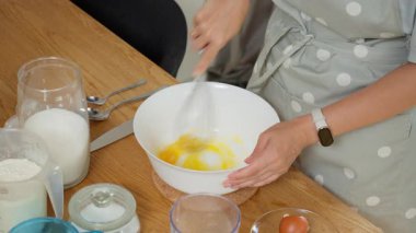 Fırına hamur hazırlamak için kasede şekerli taze tavuk yumurtası çırpıyorum. Önlüklü bir kadın mutfak masasında un ve süt kullanarak ev yapımı kek veya krep yapar..