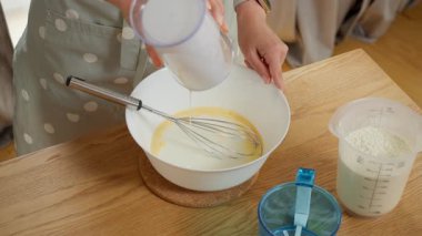 Ev yapımı hamur hamuru hazırlamak için yumurta ve un dolu bir kaseye kefir dökmek. Önlüklü kadın mutfakta yemek pişiriyor, lezzetli pasta için malzemeleri karıştırıyor ya da kahvaltıda krep yapıyor..