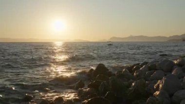 The golden sun sets over the Adriatic sea, creating a beautiful reflection on the water. Waves splash on a rocky shore in Croatia, with a small boat visible on the horizon. A romantic summer scene.