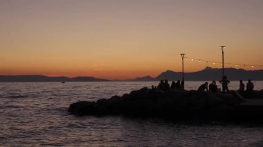 Silhouettes of people relax on a stone pier, enjoying the beautiful twilight sky over the Adriatic sea in Croatia. String lights create a warm, romantic atmosphere for a perfect summer evening.