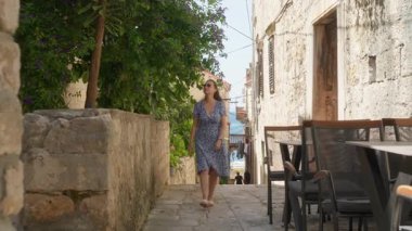 A woman in a dress is strolling through a historic city past an outdoor restaurant in Korcula. Travel lifestyle concept with sidewalk cafes, tourism, and exploring a quiet European town.