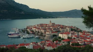 The stunning historic town of Korcula, Croatia, is revealed in a cinematic drone flight from a viewpoint on a hill, set against a backdrop of mountains and the Adriatic Sea at sunset.