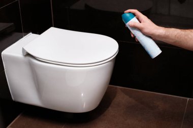 Tuvaletteki klozetin yanında oda spreyi tutan bir adamın yakın çekimi. Tuvaletteki kötü kokuyla savaşmak.