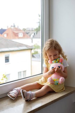 Yumuşak oyuncaklı küçük kız oturma odasının camında oturuyor.