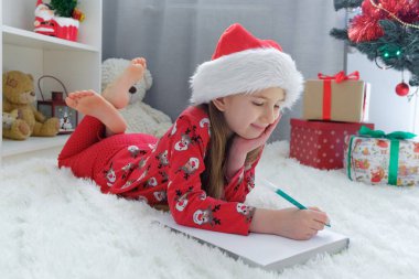 Mutlu Noeller ve mutlu Noeller. Noel ağacının altında Noel Baba şapkası giymiş mutlu bir kız Noel Baba 'ya mektup yazıyor..
