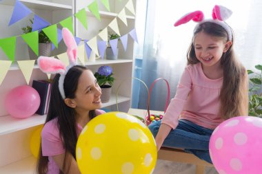 Pembe tişört ve tavşan kulağı giymiş iki kız kardeş Paskalya bayramını balonlarla süslenmiş bir odada bir sepette yumurta topluyor..
