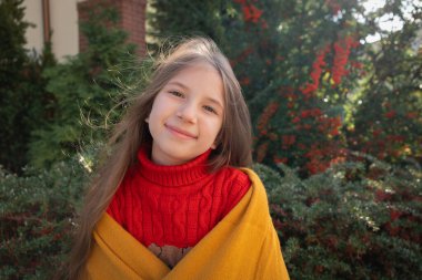Ilık sonbahar. 7-9 yaşında, kahverengi saçlı, kırmızı kazaklı ve sarı atkılı bir kız. Portakal meyveli büyük bir çalılığın yanında duruyor..
