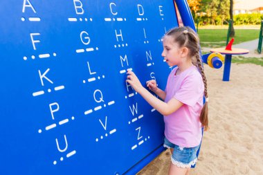 A little girl 7-8 years old with long hair dressed in a pink T-shirt and shorts is studying Morse code on a blackboard in the playground.