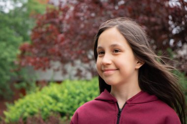 10-11 yaşındaki neşeli esmer bir kızın portresi. Uzun saçlı. Sokakta kırmızı yeşil yapraklar var..