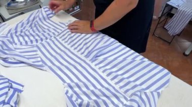 Bir insan beyaz bir masada çizgili bir gömleği ustalıkla ütüler. Odak noktası kırışıklıkları kaldırmak için kullanılan hassasiyet ve teknik, demirin kumaşın üzerinde süzülürken dikkatli hareketlerini gösteriyor.