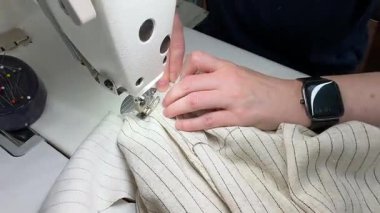 Profesyonel dikiş makinesi kullanarak ince çizgili keten kumaş diken ellerin yakın çekimi. Bir insan endüstriyel dikiş makinesine dikiş dikiyor, malzemeyi iki eliyle dikkatlice yönlendiriyor..