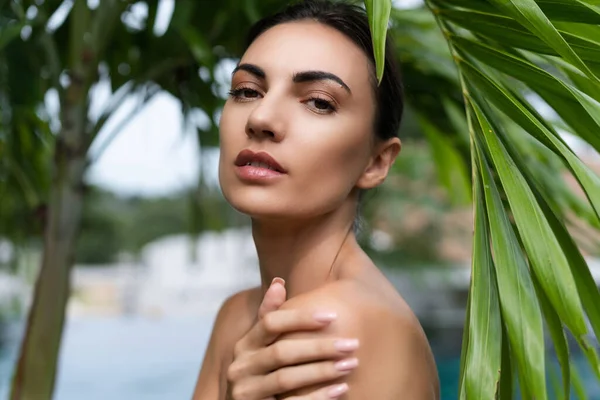 Beauty portrait, soft skin. Female model with natural make-up and healthy skin behind a green leafy plant. Portrait of a beautiful girl with nude nails, bare shoulders