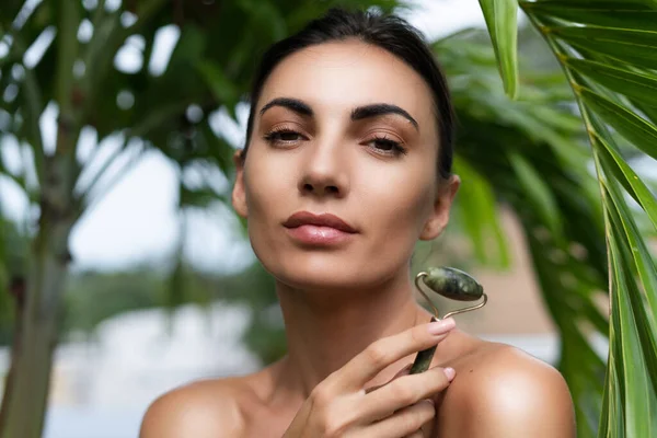 Beauty portrait, soft skin. Female model with natural make-up and healthy skin behind a green leafy plant. Portrait of a beautiful girl with nude nails, bare shoulders holding roller massager outdoor