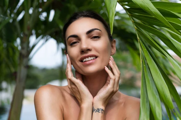 Beauty portrait, soft skin. Female model with natural make-up and healthy skin behind a green leafy plant. Portrait of a beautiful girl with nude nails, bare shoulders