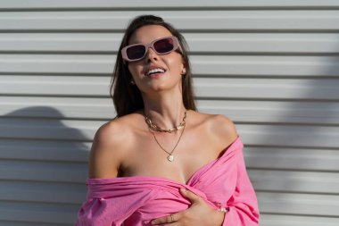 Young beautiful brunette in a pink shirt, neck jewelry, necklace, trendy sunglasses on the background of a light garage door fence