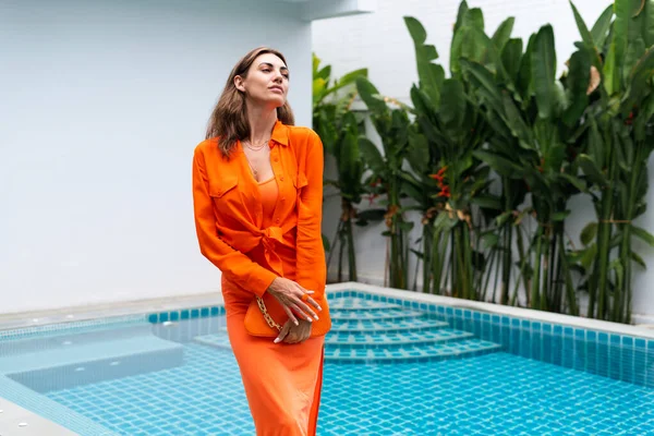 Stylish fit fashion women in bright orange fitting dress and shirt holding fashion bag posing at luxury tropical villa by pool outdoor natural day light