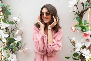 Young beautiful smiling cute romantic woman in trendy summer pink dress. Carefree woman posing in the street near white wall with flowers. Positive model outdoors in sunglasses.