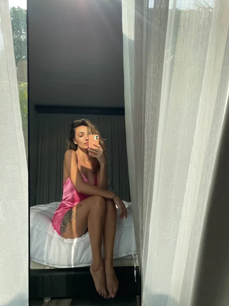 フィット日焼けした女性でピンクシルクショートスリープドレスで自宅取る写真自撮りで電話でミラーのためのソーシャルメディア — ストック写真