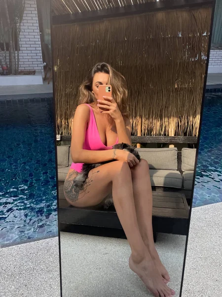在户外 穿着亮晶晶粉色泳衣的体态丰满的 晒得发亮的女人在户外的社交媒体 游泳池边与小狗垂直拍摄自己的照片 — 图库照片