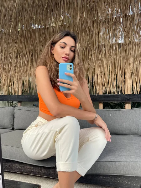 在户外 穿着橙色上衣和舒适舒适的裤子的晒黑的女人在手机上拍照 在镜子里为社交媒体 垂直拍摄 — 图库照片
