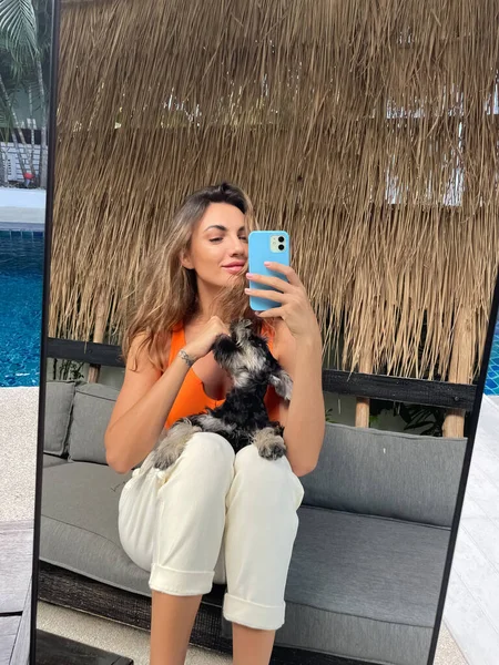 在户外 穿着橙色上衣和舒适舒适的裤子的女人可以在手机上自拍 在社交媒体 垂直镜头中还可以看到小狗的缩影 — 图库照片